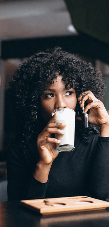 A black woman on the phone drinking a latte in a glass coffee cup