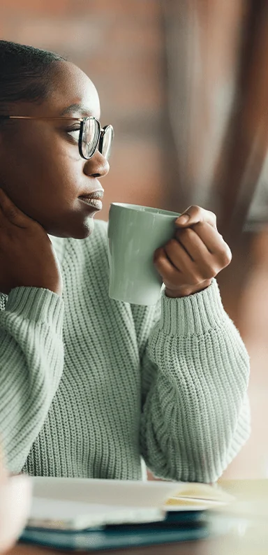 Pensive black creative woman drinking coffee while working at home office. The view is through glass.