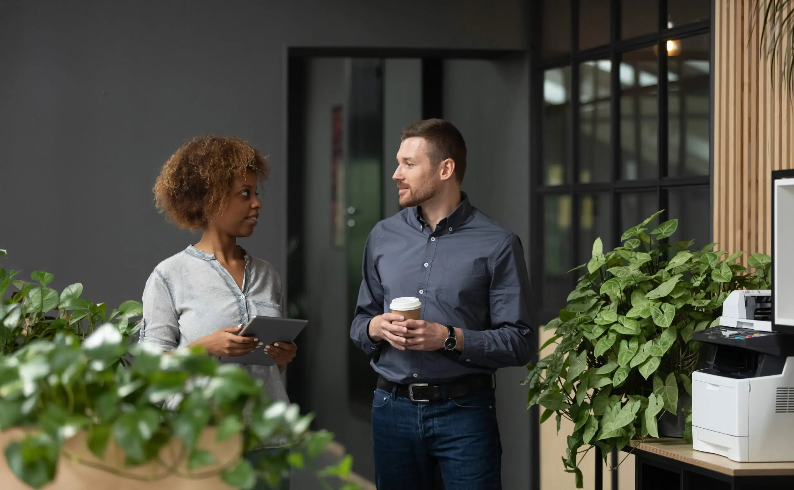 A man and woman standing by an office printer holding a cup of takeaway coffee