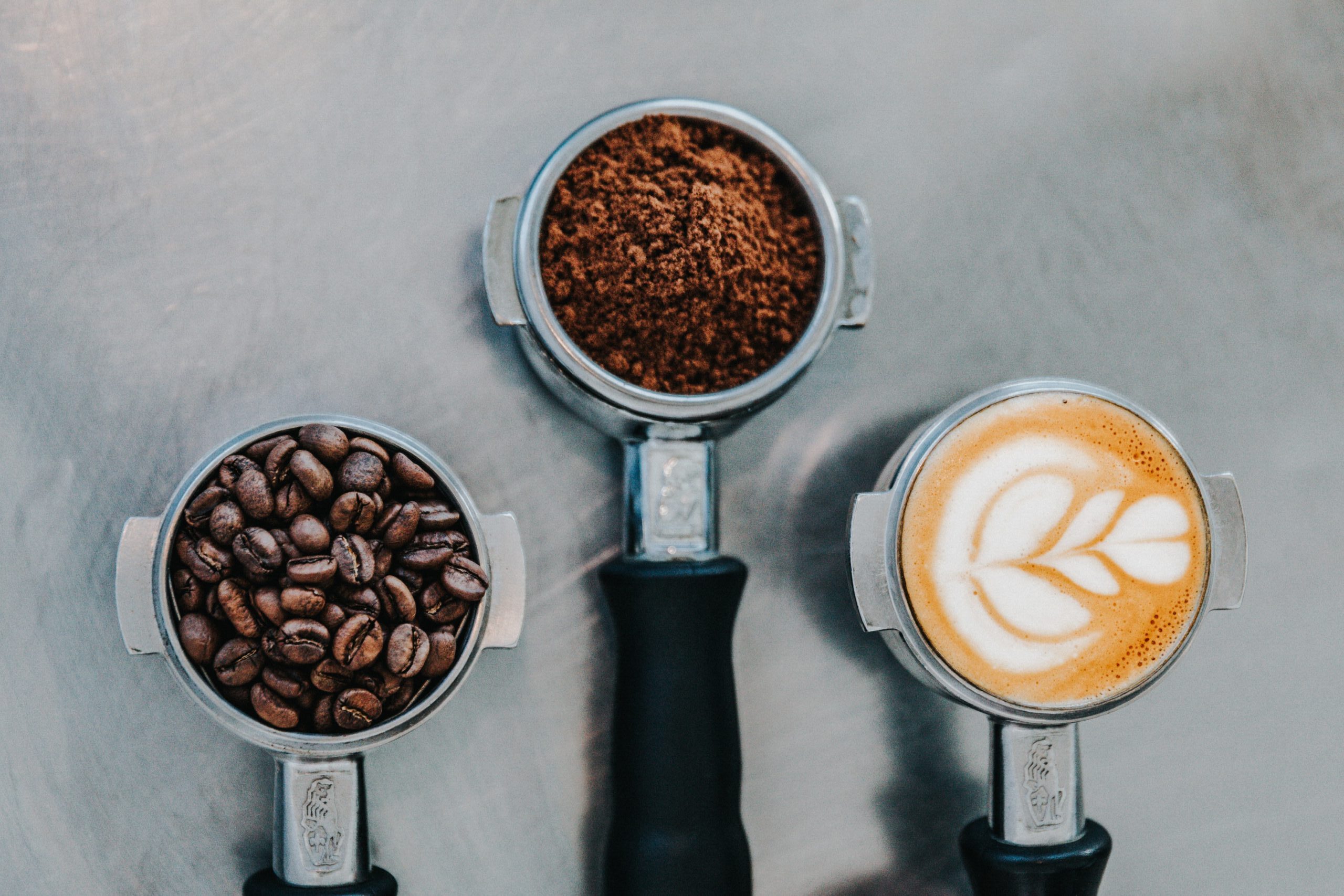 Three Commercial Espresso Coffee Machine Portafilters Filled With Coffee Beans, Ground Coffee and a Latte Coffee