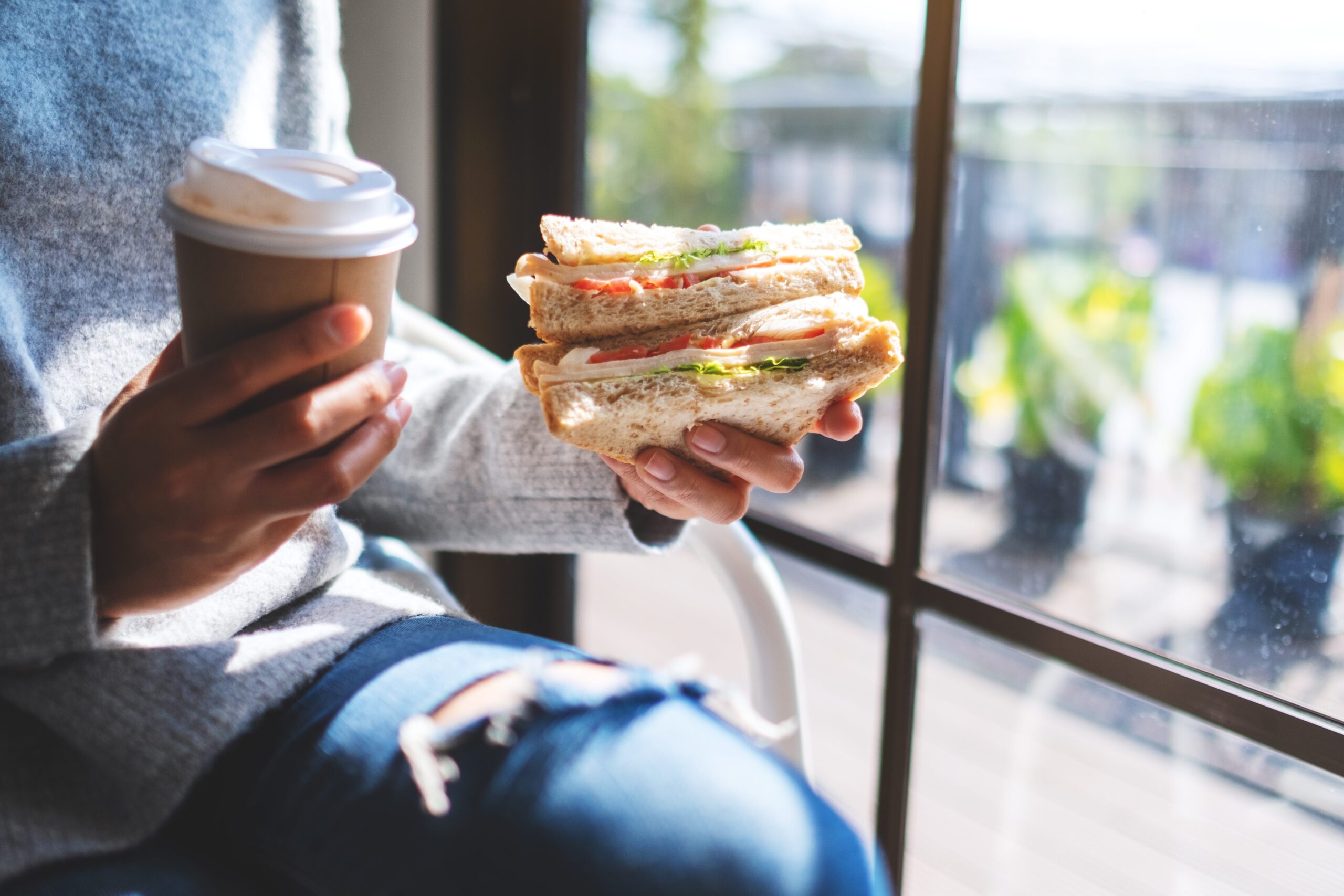 A person holding their sandwich and takeaway coffee in a recyclable container in front of a window