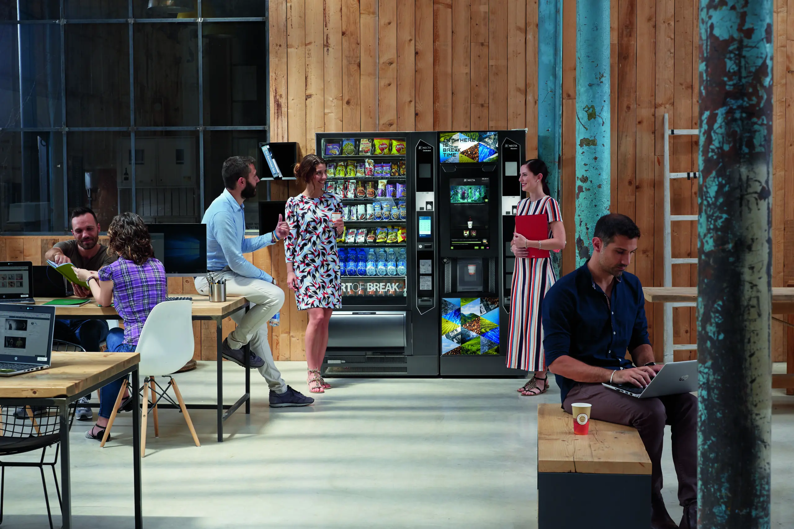 A diverse office setting with workers on a laptop or gathered around the commercial snack and drink vending machine talking.