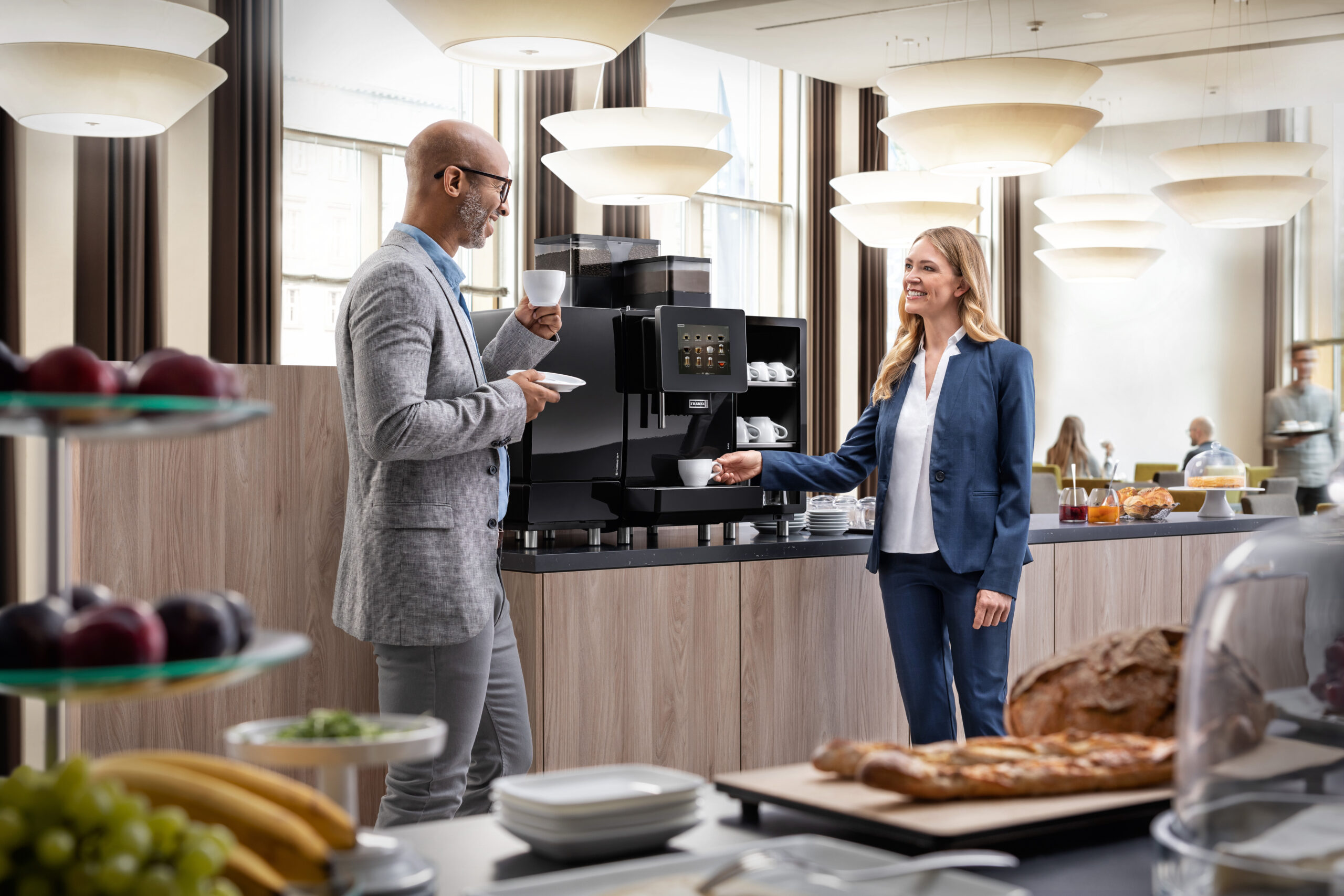 A hotel scene with a Franke A600 commercial coffee machine featuring a man and woman grabbing a cup of coffee from the machine in a porcelain cup and saucer