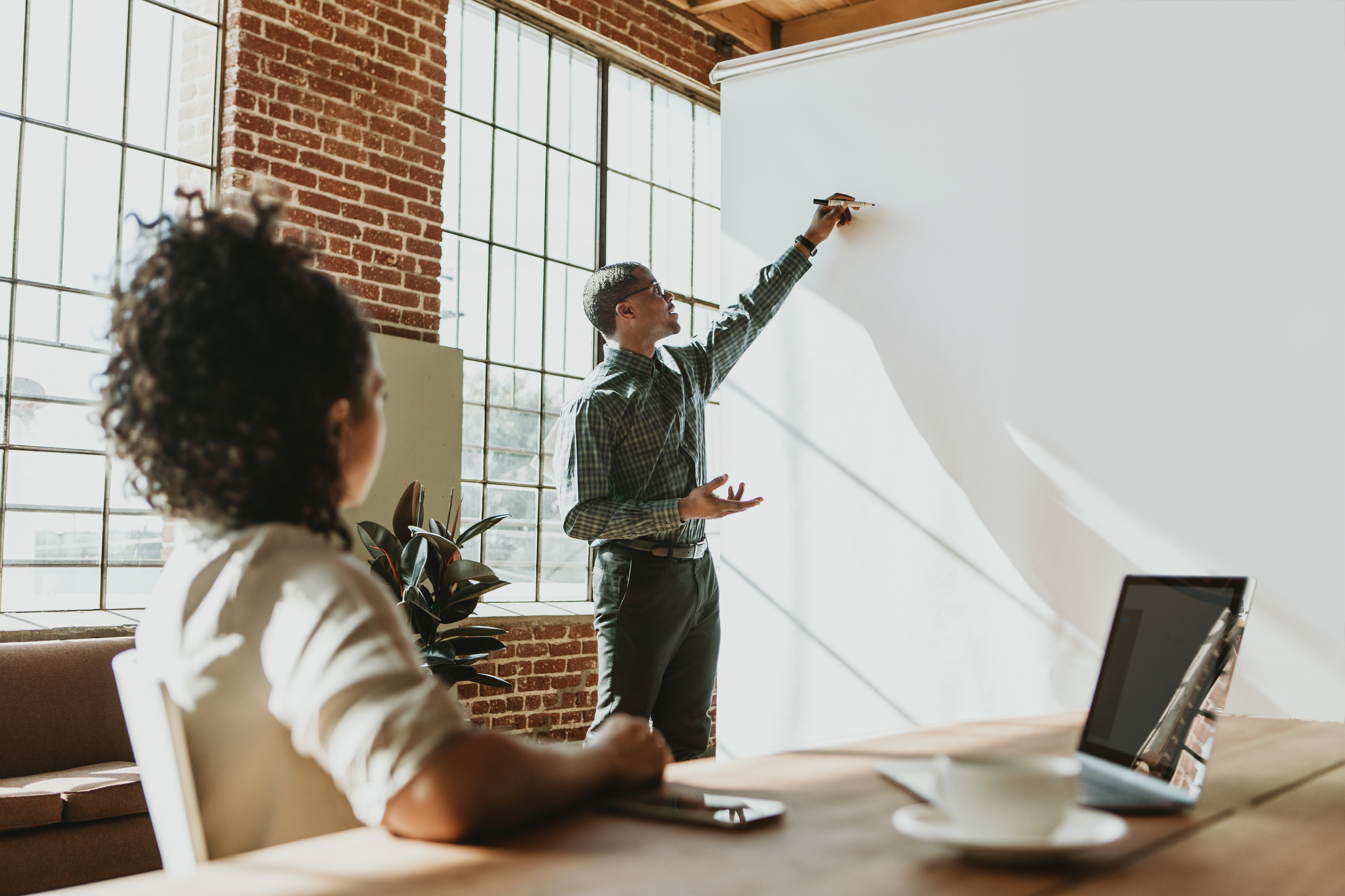 Two people sat in an office meeting room. One writing on a whiteboard and the other listening.