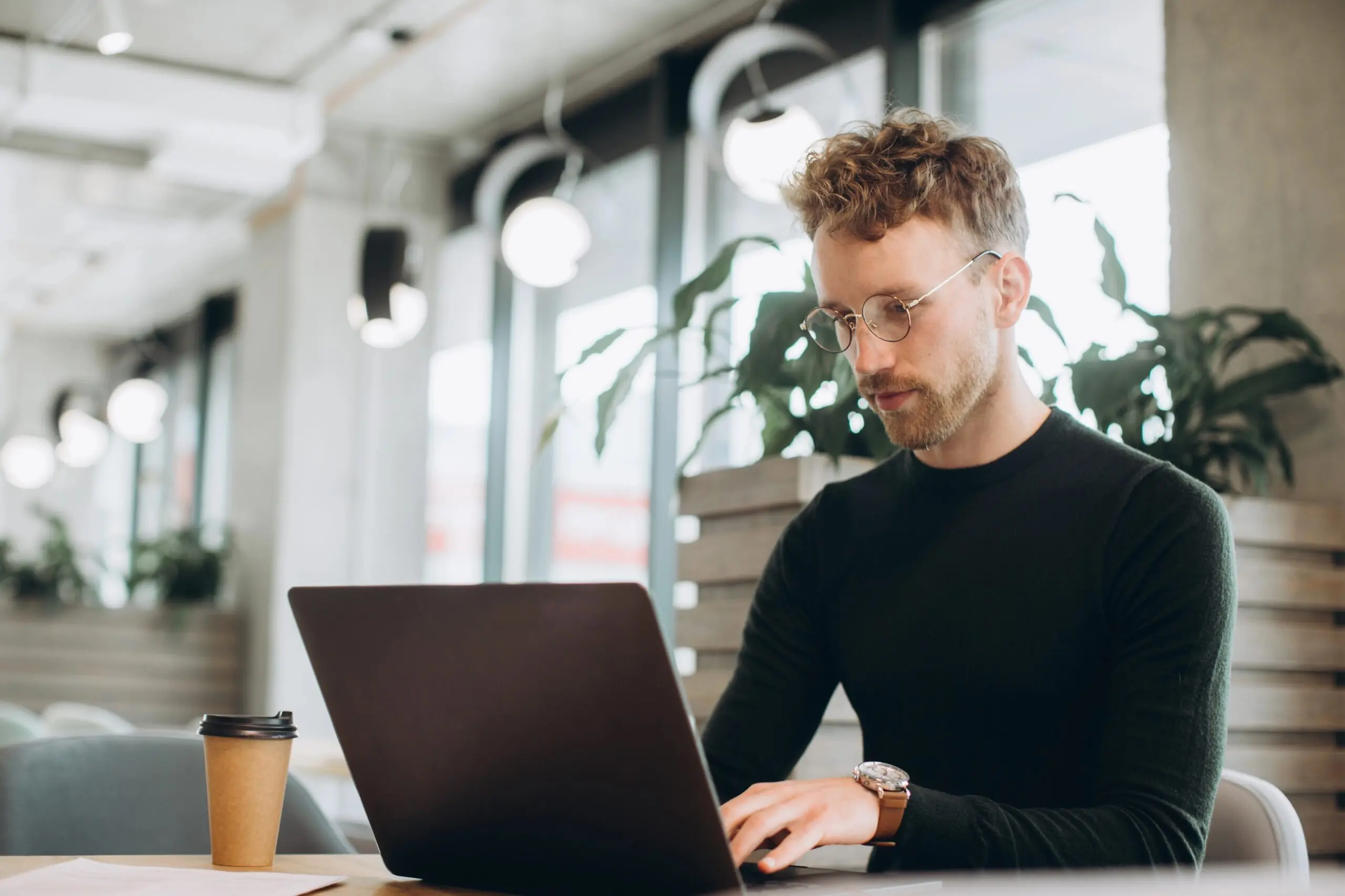 A man dressed in black working at his desk with a laptop and takeaway recyclable coffee cup