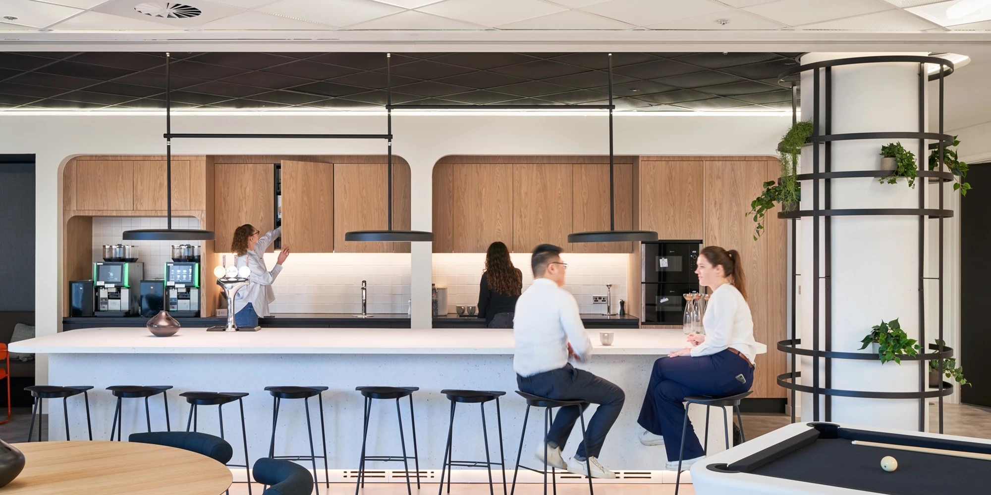 An office breakout space with people sat at a breakfast bar talking, an office coffee machine and people using the cupboards and worktops for meal or drink preparation