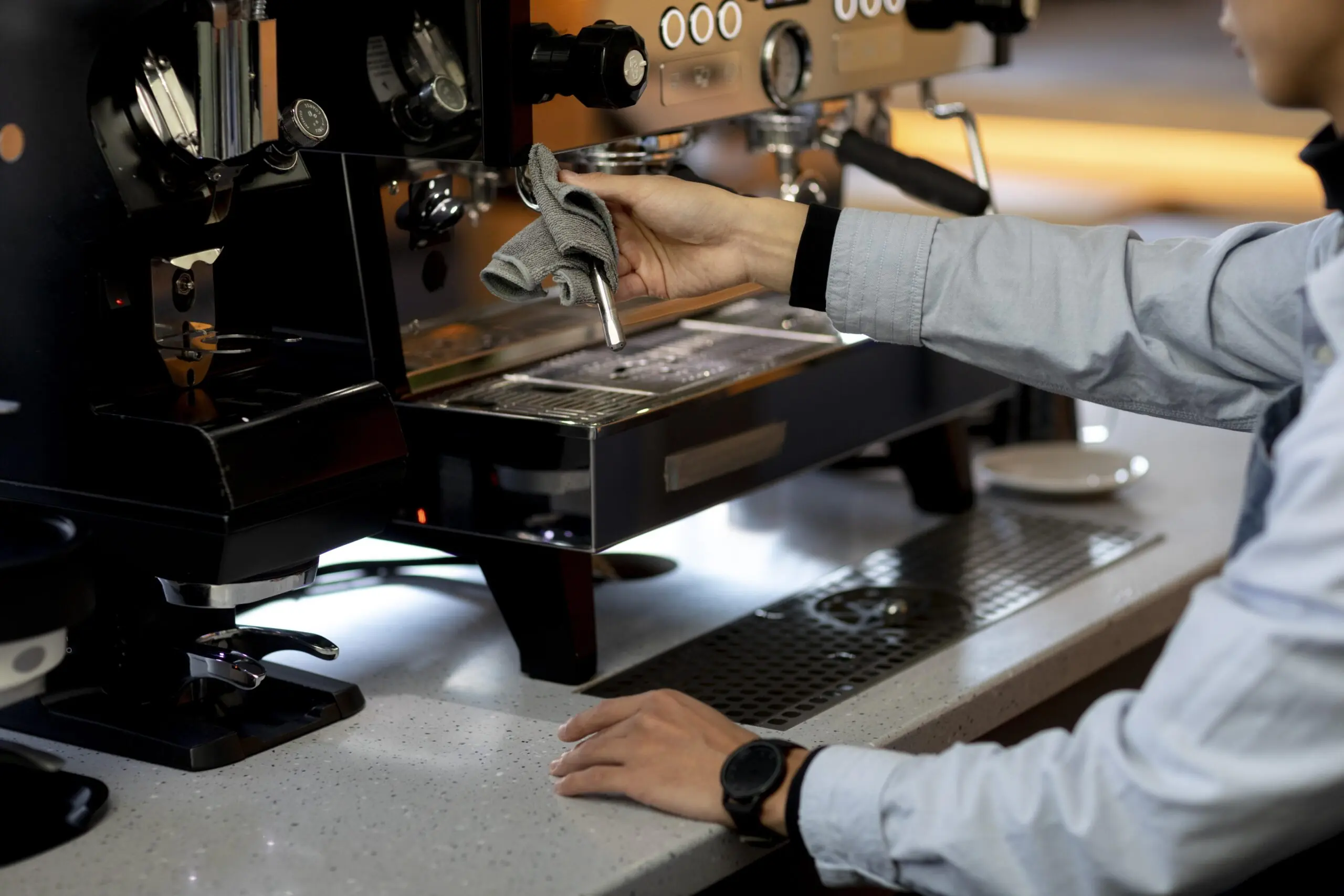 A barista staff member cleaning a commercial espresso coffee machine's steam wand with a cloth