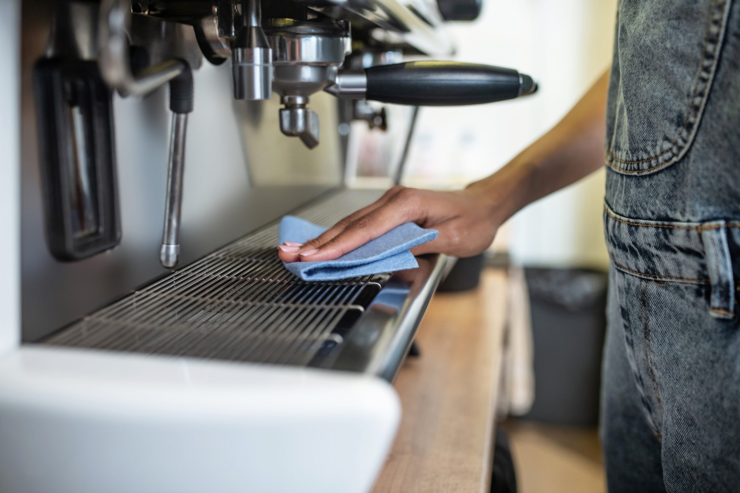 A person in dungarees cleaning a commercial espresso coffee machine with a cloth