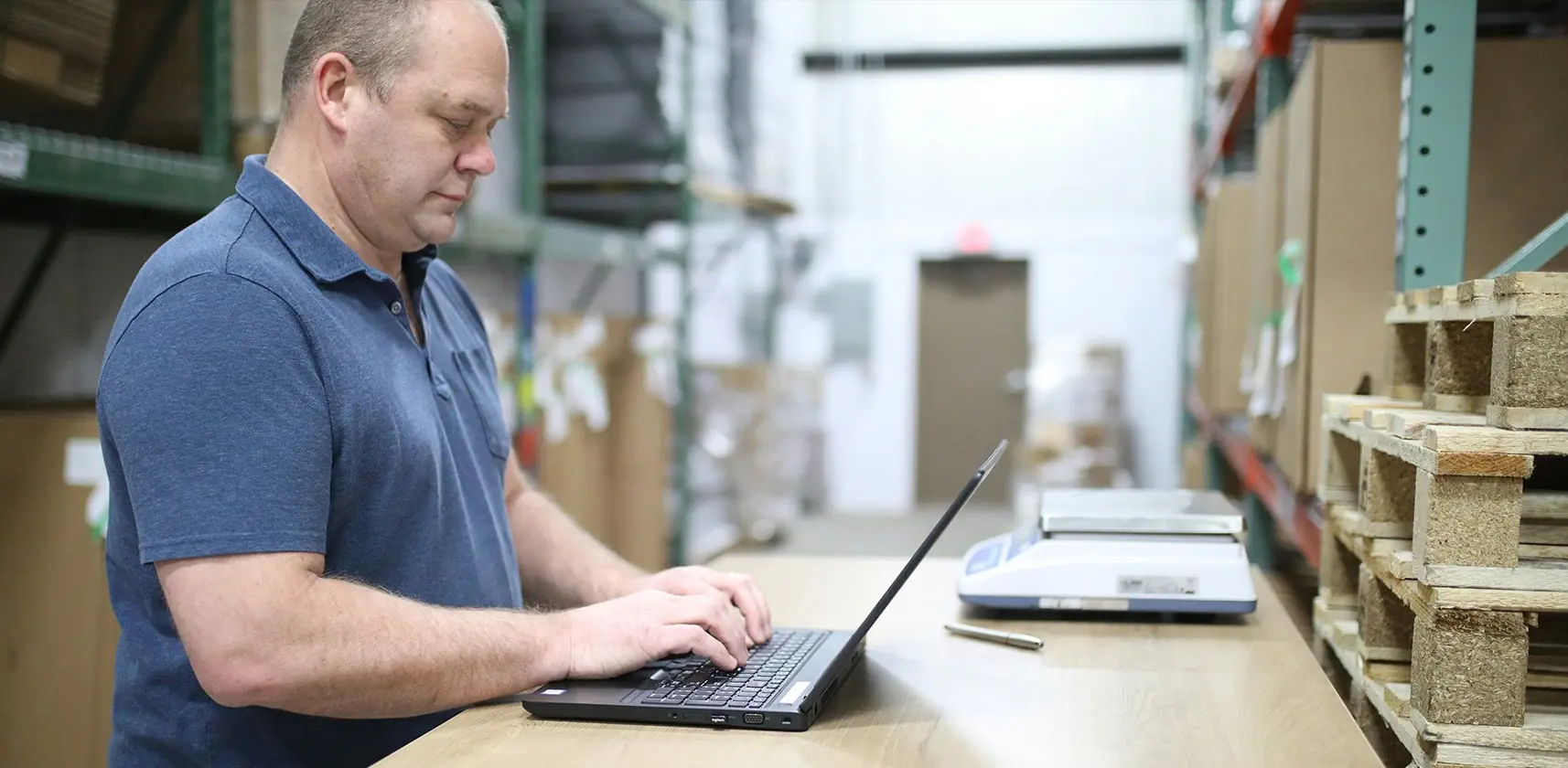 A man working in a manufacturing and distribution centre warehouse on a laptop